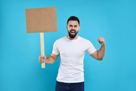 Angry man holding blank sign on light blue background, space for text