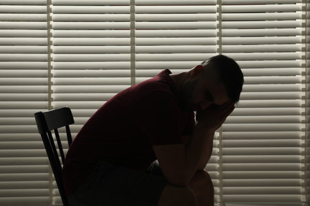 Sad man sitting near closed blinds indoorsの写真素材