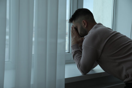 Sad man near window at home. Space for textの写真素材