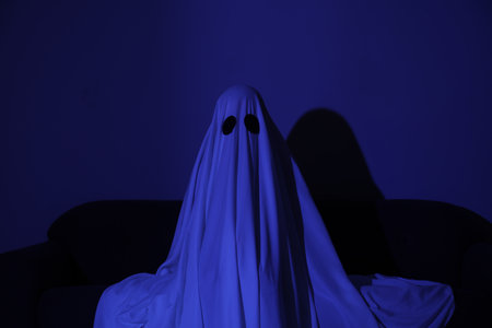 Creepy ghost. Woman covered with sheet sitting on sofa in blue lightの写真素材