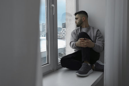 Sad man sitting on sill and looking at window indoorsの写真素材