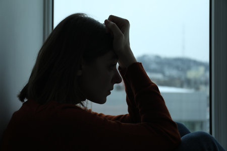 Sad young woman near window at homeの写真素材