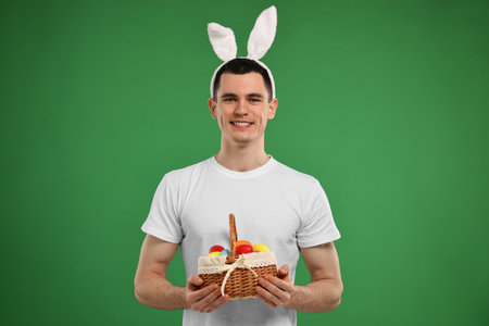 Easter celebration. Handsome young man with bunny ears holding basket of painted eggs on green...