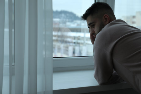 Sad man near window at home. Space for textの写真素材