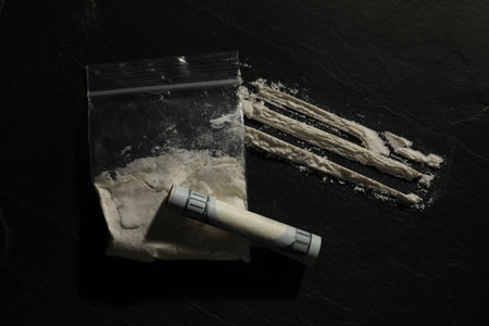 Drug addiction. Plastic bag with cocaine and rolled dollar banknote on dark textured background,...