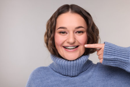 Happy woman in warm sweater pointing at braces on her teeth against gray backgroundの写真素材