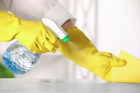 Woman with spray bottle and microfiber cloth cleaning white table at home, closeup