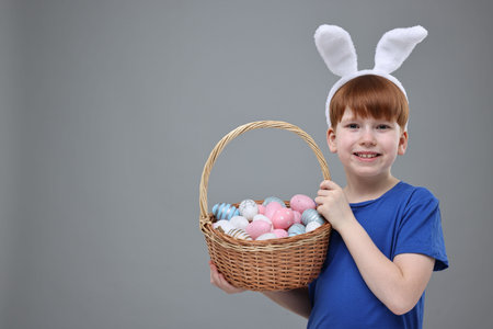 Easter celebration. Cute little boy with bunny ears and wicker basket full of painted eggs on gray...