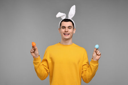 Easter celebration. Handsome young man with bunny ears holding painted eggs on gray backgroundの写真素材