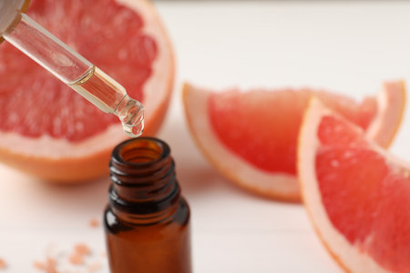 Dripping grapefruit essential oil from pipette into bottle and fresh fruit on white table, closeup. Space for textの写真素材