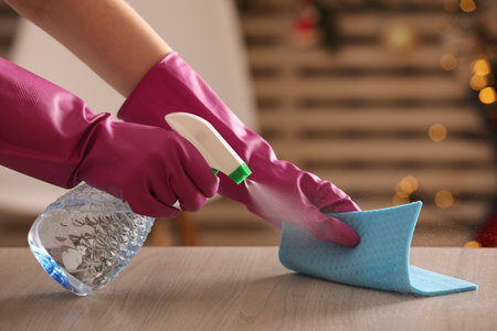 Woman with spray bottle and microfiber cloth cleaning wooden table indoors, closeup
