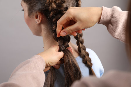 Professional stylist braiding woman's hair on gray background, closeup