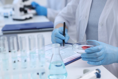 Laboratory worker holding petri dish with blood sample while working at white table, closeupの写真素材
