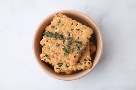 Cereal crackers with flax, sesame seeds and thyme in bowl on white marble table, top view