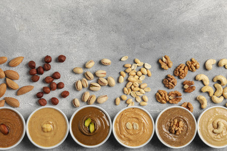 Tasty nut butters in bowls and raw nuts on light gray background, flat lay
