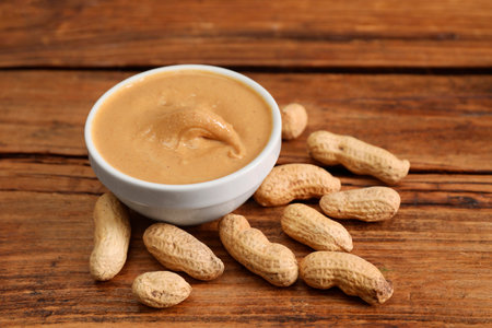 Delicious nut butter in bowl and peanuts on wooden table, closeup
