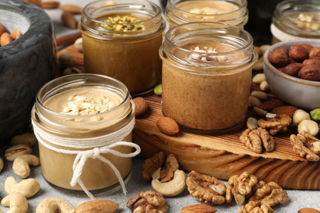 Tasty nut butters in jars and raw nuts on light gray table