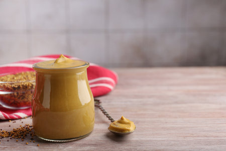 Jar and spoon with tasty mustard sauce on wooden table, space for text