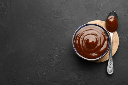 Tasty barbeque sauce in bowl and spoon on dark textured table, top view. Space for text
