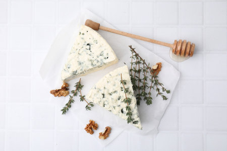Tasty blue cheese with thyme, walnuts and honey dipper on white tiled table, flat lay