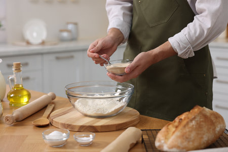 Making bread. Man putting dry yeast into bowl with flour at wooden table in kitchen, closeup