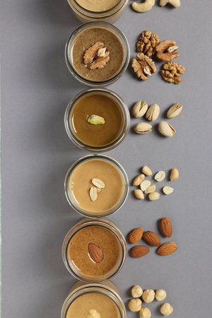 Tasty nut butters in jars and raw nuts on gray table, flat lay