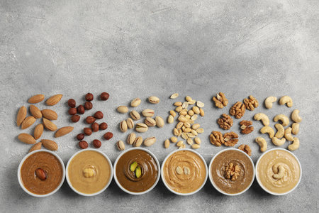 Tasty nut butters in bowls and raw nuts on light gray background, flat lay