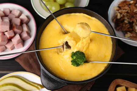 Dipping different products into fondue pot with melted cheese on black wooden table, flat layの写真素材