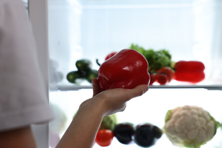 Young woman taking red bell pepper out of refrigerator at night, closeupの写真素材