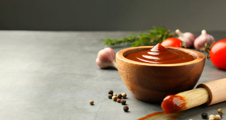 Tasty barbeque sauce in bowl, brush and peppercorns on gray textured table, closeup. Space for textの写真素材
