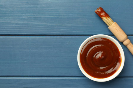Tasty barbeque sauce in bowl and brush on blue wooden table, top view. Space for textの写真素材