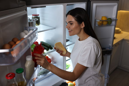 Young woman taking ketchup and cheese out of refrigerator in kitchen at nightの写真素材