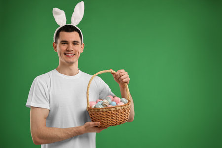 Easter celebration. Handsome young man with bunny ears holding basket of painted eggs on green background. Space for textの写真素材