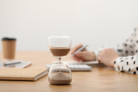 Hourglass with flowing sand on desk. Woman taking notes while using calculator indoors, selective...