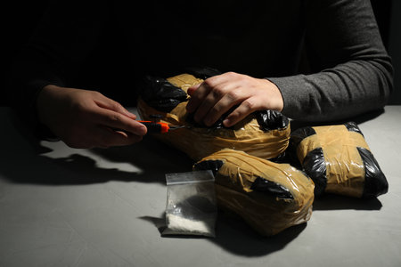 Smuggling and drug trafficking. Man opening package of narcotics with box cutter in darkness,...