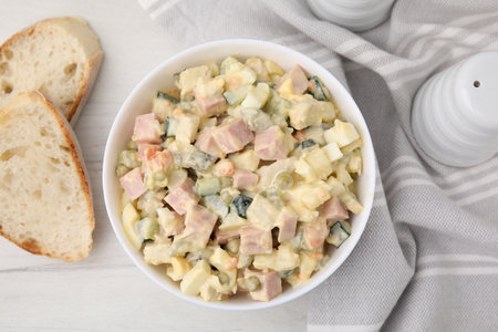 Tasty Olivier salad with boiled sausage in bowl and bread on white wooden table, flat layの写真素材