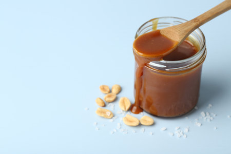 Jar of tasty salted caramel, spoon and peanuts on light blue background, closeup. Space for textの写真素材