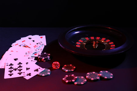 Roulette wheel with ball, chips, dice and playing cards on dark table, closeup. Casino game
