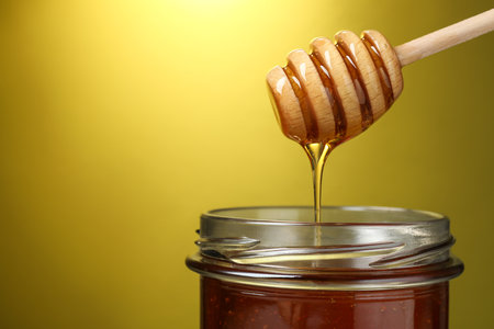 Pouring tasty honey from dipper into glass jar on golden background, closeup. Space for textの写真素材