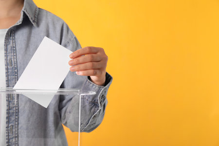 Woman putting her vote into ballot box on orange background, closeup. Space for textの写真素材