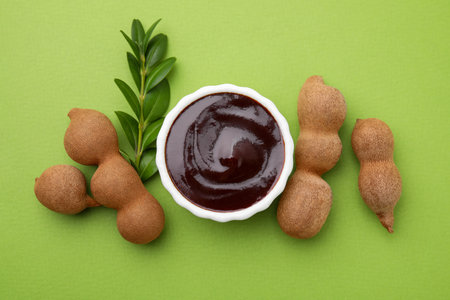 Bowl of tasty tamarind sauce, leaves and fresh pods on light green table, flat layの写真素材