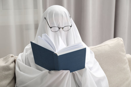 Creepy ghost. Person covered with white sheet reading book on sofa at homeの写真素材