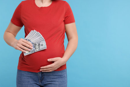 Surrogate mother. Pregnant woman with dollar banknotes on light blue background, closeup. Space for textの写真素材