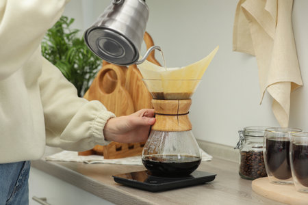 Woman pouring hot water into glass coffeemaker with paper filter and coffee at countertop in...