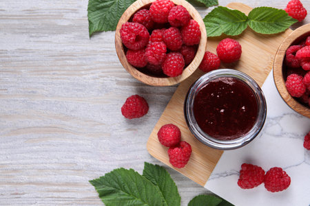 Delicious raspberry jam, fresh berries and green leaves on light wooden table, flat lay. Space for textの写真素材