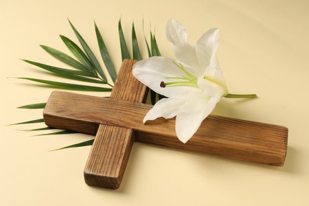 Wooden cross, lily flower and palm leaf on pale yellow background. Easter attributesの写真素材