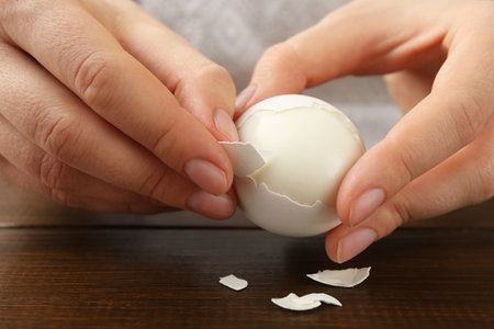 Woman peeling fresh boiled egg at wooden table, closeup