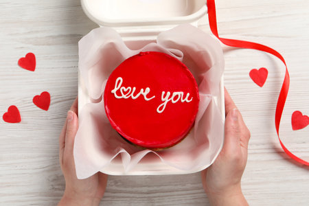 Woman holding takeaway box with bento cake at white wooden table, top view. St. Valentine's day...