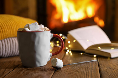 Mug with hot cocoa, marshmallows, lights and book on wooden table near fireplaceの写真素材