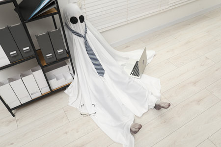 Overworked ghost. Man in white sheet with laptop on floor in office, above viewの写真素材